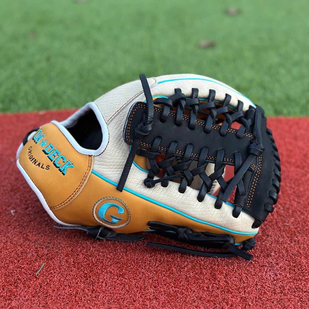 A close-up of a custom baseball glove resting on a red textured surface, likely an athletic field. The glove is primarily tan with black lacing and blue accents. On the thumb section, there is an embroidered patch with the letter “G” in a stylized font inside a blue circle. The intricate lacing and stitching are clearly visible, showcasing the craftsmanship of the glove.