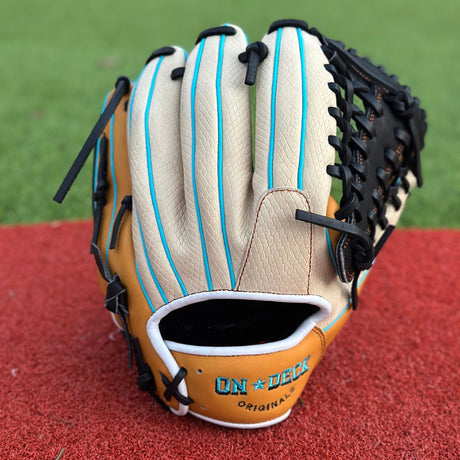 A close-up of a custom baseball glove resting on a red textured surface, likely an athletic field. The glove is primarily tan with black lacing and blue accents. On the thumb section, there is an embroidered patch with the letter “G” in a stylized font inside a blue circle. The intricate lacing and stitching are clearly visible, showcasing the craftsmanship of the glove.