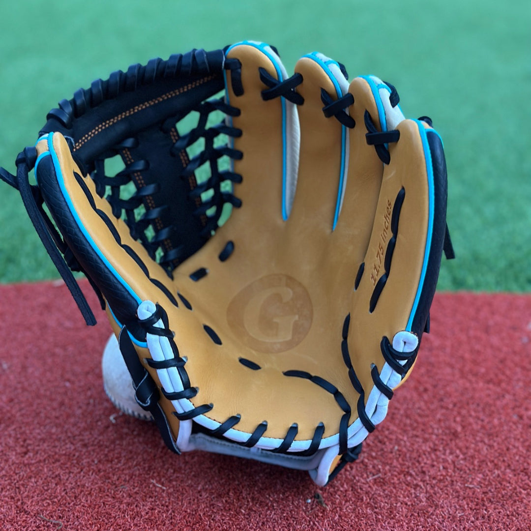 A close-up of a custom baseball glove resting on a red textured surface, likely an athletic field. The glove is primarily tan with black lacing and blue accents. On the thumb section, there is an embroidered patch with the letter “G” in a stylized font inside a blue circle. The intricate lacing and stitching are clearly visible, showcasing the craftsmanship of the glove.