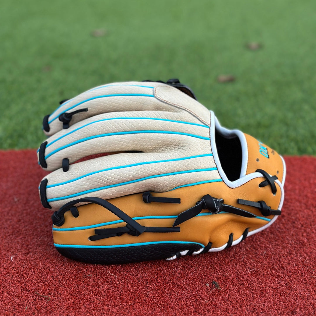 A close-up of a custom baseball glove resting on a red textured surface, likely an athletic field. The glove is primarily tan with black lacing and blue accents. On the thumb section, there is an embroidered patch with the letter “G” in a stylized font inside a blue circle. The intricate lacing and stitching are clearly visible, showcasing the craftsmanship of the glove.