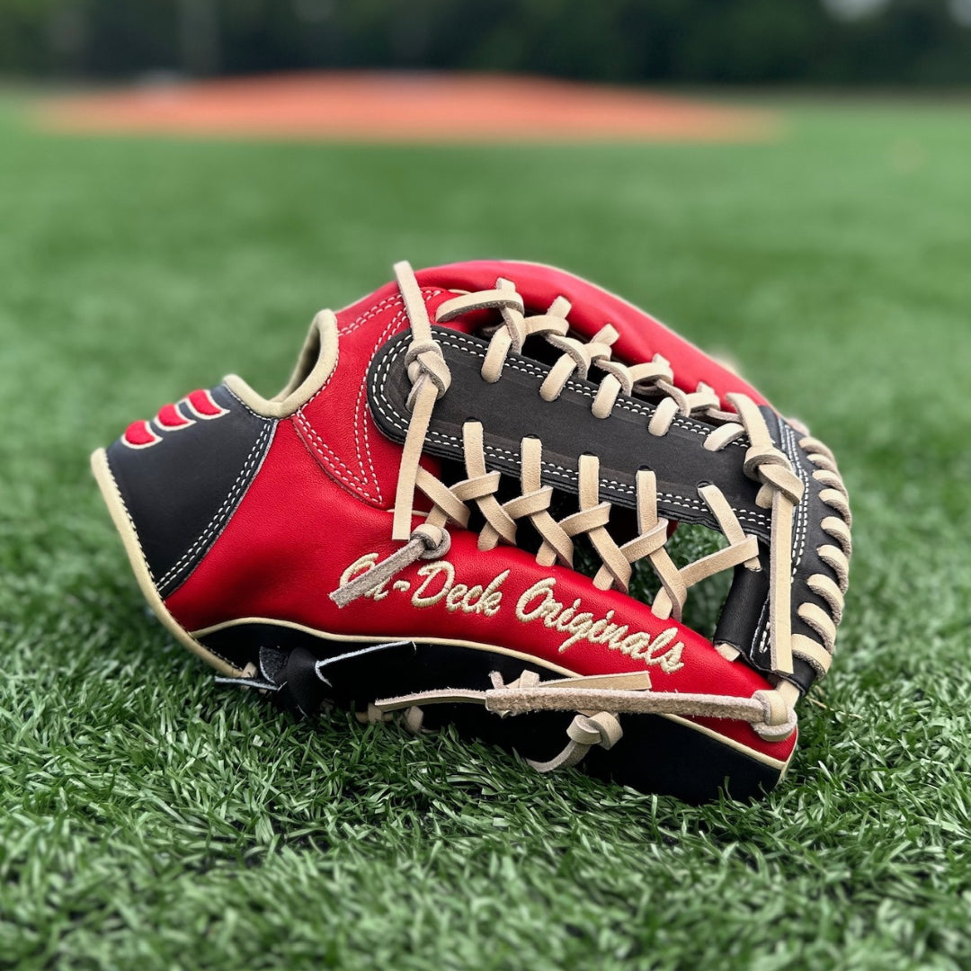 A close-up image of a red and black baseball glove resting on a grassy field. The glove is positioned with the palm facing down, showcasing its intricate blonde lacing, padded leather design, and text ‘On-Deck Originals’ on the thumb. In the background, there is a blurred view of a baseball diamond, indicating that the setting is likely a sports field. The focus on the glove against the soft-focus backdrop highlights its details and suggests its readiness for use in a game.